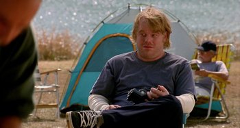 Movie still from “Love Liza” (2002), directed by Todd Louiso – A man sitting in front of a tent holding a camera; Medium shot, Over the shoulder angle