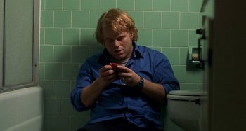 Movie still from “Love Liza” (2002), directed by Todd Louiso – A man sitting on a toilet looking at his cell phone; Medium shot, High angle