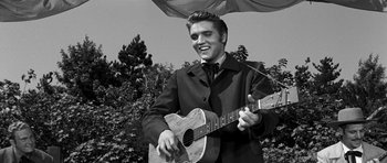 Movie still from “Love Me Tender” (1956), directed by Robert D. Webb – A man in a suit and tie playing a guitar in front of trees; Medium shot, Low angle