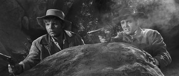 Movie still from “Love Me Tender” (1956), directed by Robert D. Webb – Two men in hats are pointing a gun at a rock; Medium shot, Low angle
