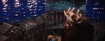 A man with a hat on holding paintbrushes and a painting
