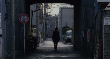 Movie still from “Maborosi” (1995), directed by Kore-eda Hirokazu – A person walking down a street under an overpass; Wide shot, Over the shoulder angle