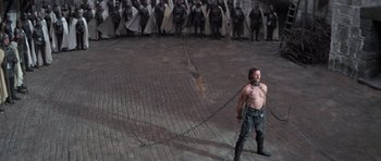 Movie still from “Macbeth” (1971), directed by Roman Polanski – A man chained up to a chain standing in front of a group of people; Wide shot, High angle