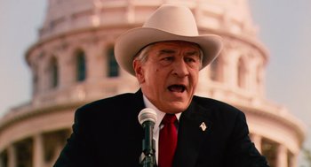 Movie still from “Machete” (2010), directed by Robert Rodriguez – An older man wearing a white cowboy hat and speaking into a microphone in front of a building; Close Up shot, Over the shoulder angle