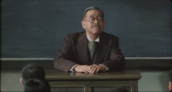 Movie still from “Madadayo” (1993), directed by Akira Kurosawa – An older man sitting at a table in front of a chalkboard; Medium shot, Low angle