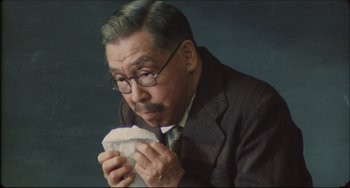 Movie still from “Madadayo” (1993), directed by Akira Kurosawa – An older man in a suit and glasses eating a sandwich; Close Up shot, Low angle