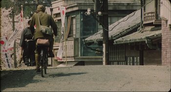 Movie still from “Madadayo” (1993), directed by Akira Kurosawa – A man riding a bike down a street past a building; Extreme Wide shot, High angle