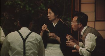 Movie still from “Madadayo” (1993), directed by Akira Kurosawa – A woman eating food while sitting next to two other people; Medium shot, Over the shoulder angle
