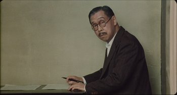 Movie still from “Madadayo” (1993), directed by Akira Kurosawa – An older man sitting at a table with a pen in his hand; Medium shot, Low angle