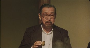 Movie still from “Madadayo” (1993), directed by Akira Kurosawa – An older man wearing glasses and a brown jacket holding a donut; Medium shot, Low angle
