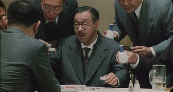 Movie still from “Madadayo” (1993), directed by Akira Kurosawa – An older man sitting at a dinner table surrounded by other men; Medium shot, High angle