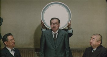 Movie still from “Madadayo” (1993), directed by Akira Kurosawa – A man in a suit and tie holding up two fingers; Medium shot, Over the shoulder angle