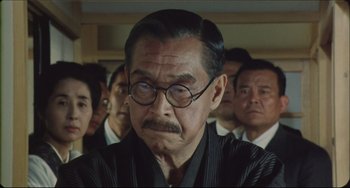 Movie still from “Madadayo” (1993), directed by Akira Kurosawa – An older man with glasses is surrounded by a group of people; Close Up shot, Over the shoulder angle