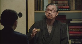 Movie still from “Madadayo” (1993), directed by Akira Kurosawa – An older man wearing a black robe and holding a piece of food; Medium shot, Over the shoulder angle
