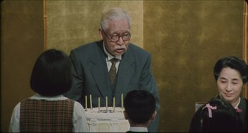 Movie still from “Madadayo” (1993), directed by Akira Kurosawa – An old man in a suit stands in front of a birthday cake; Medium shot, Over the shoulder angle