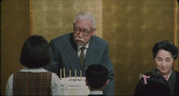 Movie still from “Madadayo” (1993), directed by Akira Kurosawa – An old man in a suit and glasses stands next to a birthday cake; Medium shot, Over the shoulder angle