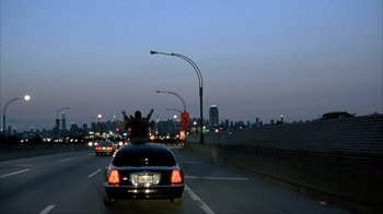 Movie still from “Made” (2001), directed by Jon Favreau – A car driving down a street at night with a person on the back of the car; Extreme Wide shot, Low angle