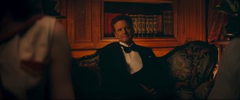 Movie still from “Magic in the Moonlight” (2014), directed by Woody Allen – A man in a tuxedo sitting on a couch in front of a book case; Medium shot, Low angle