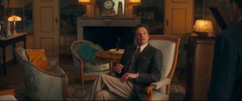 Movie still from “Magic in the Moonlight” (2014), directed by Woody Allen – A man in a suit sitting in a chair in front of a fireplace; Medium shot, Over the shoulder angle