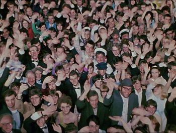 Movie still from “Magical Mystery Tour” (1967), directed by Ringo Starr – A large group of people in a room with their hands in the air; Wide shot, Overhead angle
