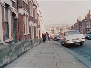 Movie still from “Magical Mystery Tour” (1967), directed by Ringo Starr – Two people walking down a street with a car parked on the side of the road; Extreme Wide shot, High angle