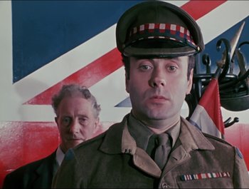 Movie still from “Magical Mystery Tour” (1967), directed by Ringo Starr – A man in a military uniform stands in front of an american flag; Medium shot, Low angle