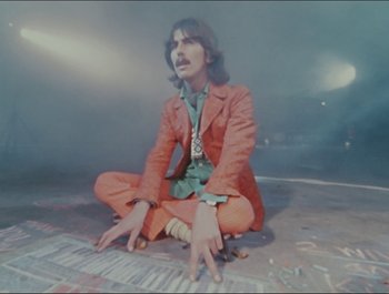 Movie still from “Magical Mystery Tour” (1967), directed by Ringo Starr – A man sitting on the ground wearing an orange suit; Medium shot, High angle