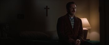 Movie still from “Magnolia” (1999), directed by Paul Thomas Anderson – A man sitting on a couch in front of a cross on the wall; Medium shot, Low angle