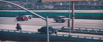 Movie still from “Magnum Force” (1973), directed by Ted Post – Cars driving down a road with a bridge in the background; Extreme Wide shot, High angle