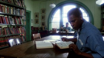 Movie still from “Malcolm X” (1992), directed by Spike Lee – A man sitting at a wooden table with a book in front of him; Medium shot, Low angle