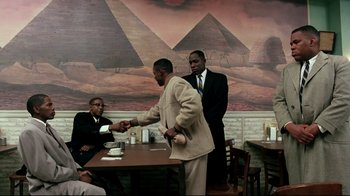 Movie still from “Malcolm X” (1992), directed by Spike Lee – Two men shaking hands at a table in front of a painting; Wide shot, Over the shoulder angle