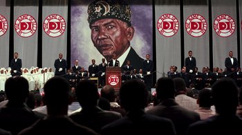 Movie still from “Malcolm X” (1992), directed by Spike Lee – A crowd of people sitting in front of a painting of an african - american man; Extreme Wide shot, High angle