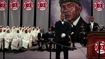 Movie still from “Malcolm X” (1992), directed by Spike Lee – A group of people in front of a painting of an african - american man; Wide shot, Low angle