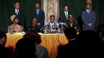 Movie still from “Malcolm X” (1992), directed by Spike Lee – A group of men sitting at a table with microphones; Medium shot, Low angle