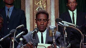 Movie still from “Malcolm X” (1992), directed by Spike Lee – A man sitting at a table with microphones; Medium shot, Low angle