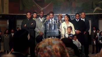 Movie still from “Malcolm X” (1992), directed by Spike Lee – A group of people standing around a man speaking at a podium; Medium shot, High angle