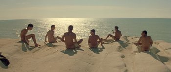 Movie still from “Malena” (2000), directed by Giuseppe Tornatore – A group of men sitting on a rock near the ocean; Wide shot, High angle