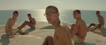 Movie still from “Malena” (2000), directed by Giuseppe Tornatore – A group of young men sitting on top of a sandy beach; Medium shot, Low angle