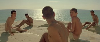 Movie still from “Malena” (2000), directed by Giuseppe Tornatore – A group of people sitting on top of a sandy beach; Medium shot, High angle