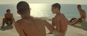 Movie still from “Malena” (2000), directed by Giuseppe Tornatore – A man and a boy sitting on the beach looking out at the ocean; Medium shot, Over the shoulder angle