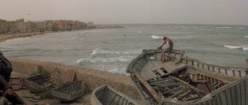 Movie still from “Malena” (2000), directed by Giuseppe Tornatore – A man standing next to an old boat on the beach; Extreme Wide shot, High angle