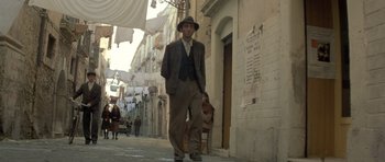 Movie still from “Malena” (2000), directed by Giuseppe Tornatore – A man wearing a suit and hat standing on the sidewalk; Wide shot, Low angle