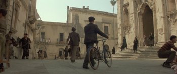 Movie still from “Malena” (2000), directed by Giuseppe Tornatore – A man holding a bicycle in front of a building; Extreme Wide shot, High angle