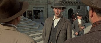 Movie still from “Malena” (2000), directed by Giuseppe Tornatore – A man wearing a suit and a hat standing in front of a building; Medium shot, Over the shoulder angle