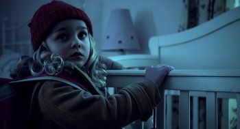 Movie still from “Mama” (2013), directed by Andy Muschietti – A little girl wearing a red hat looking over a crib; Close Up shot, High angle