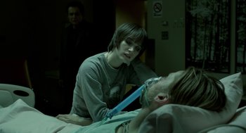 Movie still from “Mama” (2013), directed by Andy Muschietti – A man with a knife in his mouth is sitting on a hospital bed; Medium shot, Over the shoulder angle