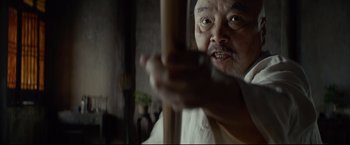 Movie still from “Man of Tai Chi” (2013), directed by Keanu Reeves – An older man holding a wooden stick in a room; Close Up shot, Low angle