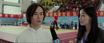 Movie still from “Man of Tai Chi” (2013), directed by Keanu Reeves – A man holding a microphone in front of an audience in an arena; Close Up shot, Low angle