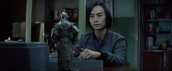Movie still from “Man of Tai Chi” (2013), directed by Keanu Reeves – A man sitting in front of a statue holding a knife; Medium shot, Over the shoulder angle