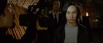 Movie still from “Man of Tai Chi” (2013), directed by Keanu Reeves – A man in grey shirt and black jacket next to a camera; Close Up shot, Over the shoulder angle